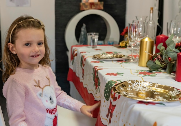 Porträt eines kleinen Mädchens, das an einem dekorierten Tisch sitzt und das Weihnachtsessen mit Blick auf die Kamera genießt