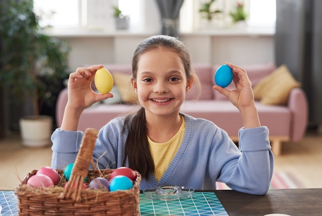 Porträt eines kleinen Mädchens, das in die Kamera lächelt, während es am Tisch sitzt und Eier für Ostern malt