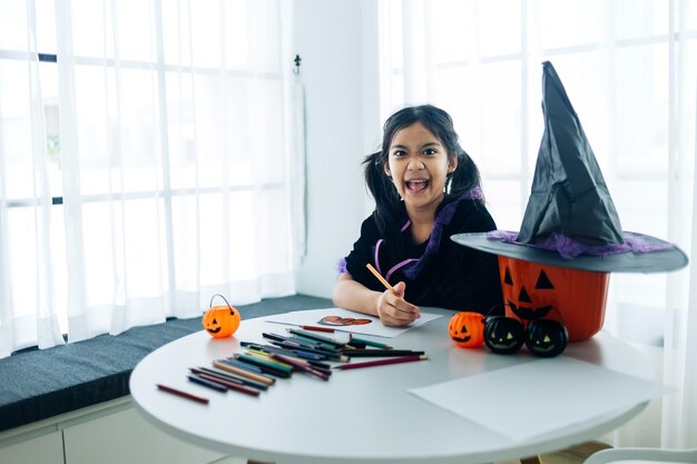 Porträt eines kleinen Mädchens im Hexenkostüm, das zu Hause Halloween-Kürbis auf den Tisch malt