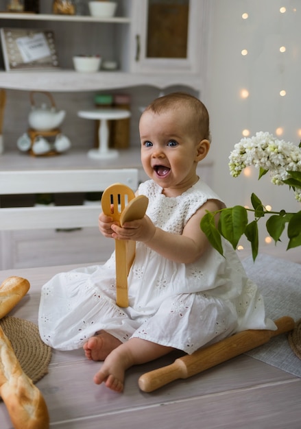 Porträt eines kleinen Mädchens in einem weißen Baumwollkleid, das auf einem Tisch sitzt und mit Holzschaufeln spielt