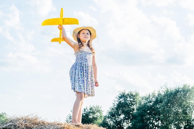 Porträt eines kleinen Mädchens in Hut und Kleid, das fliegendes gelbes Spielzeugflugzeug spielt, das auf Heuhaufen steht und in den Himmel zielt