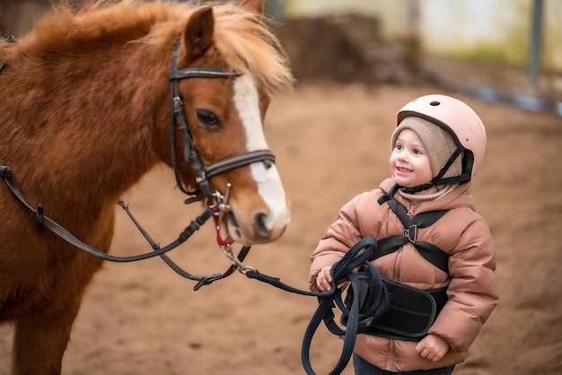 Porträt eines kleinen Mädchens in Schutzjacke und Helm mit ihrem braunen Pony vor der Reitstunde