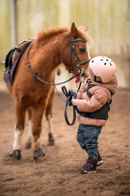 Porträt eines kleinen Mädchens in Schutzjacke und Helm mit ihrem braunen Pony vor der Reitstunde