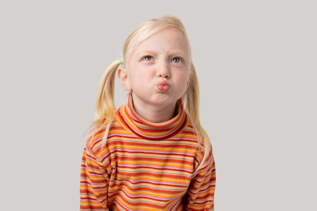 Porträt eines kleinen Mädchens mit blonden Haaren in einem gestreiften T-Shirt, das einen Kuss über einem grauen Hintergrund bläst