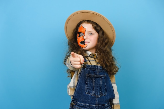 Foto porträt eines kleinen mädchens mit halloween-make-up-maske trägt hut und hemd, zeigt zeigefingerkamera auf ihre motivation, posiert auf blauem hintergrund im studio. party-urlaubskonzept