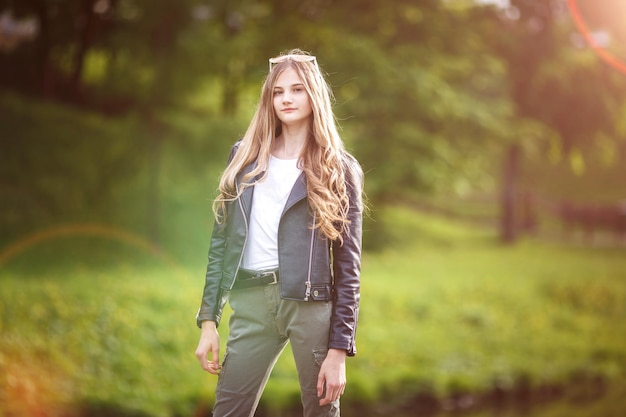 Porträt eines kleinen schönen, stilvollen, großen Mädchens mit langen Haaren in schwarzer Lederjacke im Stadtpark auf grünem Waldhintergrund