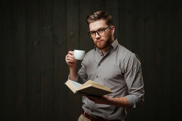 Porträt eines konzentrierten bärtigen Mannes, der ein offenes Buch hält und Kaffee trinkt, isoliert auf einer schwarzen Holzoberfläche