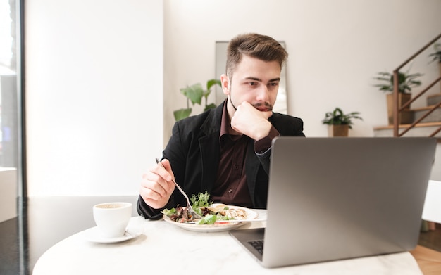 Porträt eines konzentrierten Mannes, der an einem Tisch in einem Café mit einem Teller Salat und einer Tasse Kaffee sitzt und einen Laptop benutzt.