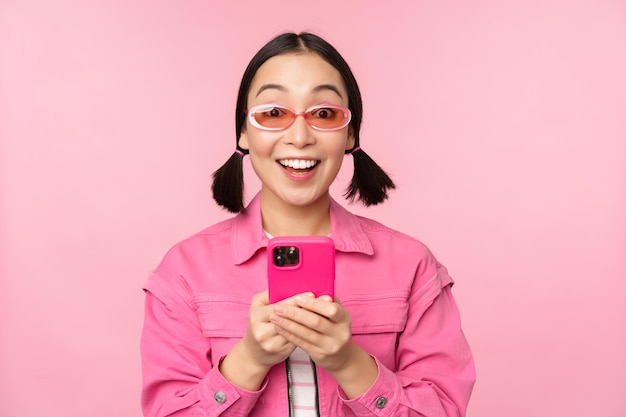 Porträt eines koreanischen Mädchens mit stylischer Sonnenbrille, das ein Handy mit einer Smartphone-App hält, die über einem rosa Hintergrund steht