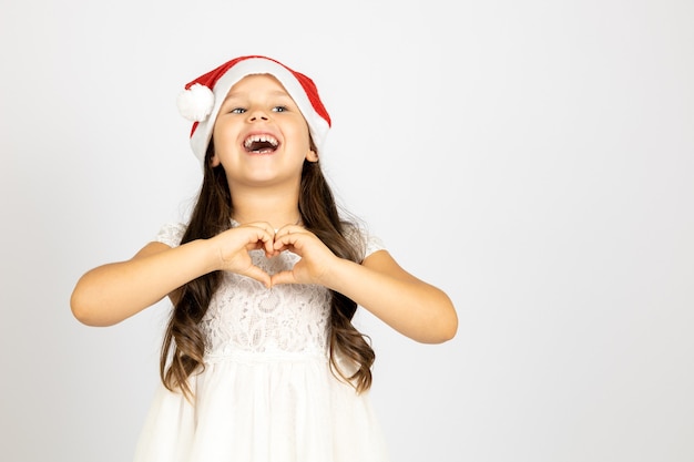 Porträt eines lachenden fröhlichen Mädchens mit langen Locken im weißen Kleid mit rotem Weihnachtsmann-Hut zeigen h...