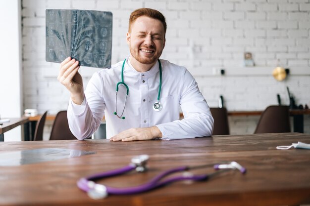 Porträt eines lachenden männlichen Arztes in weißer Uniform, der den Computertomographie-Scan des Gehirns untersucht, der am Schreibtisch im Krankenhausbüro sitzt