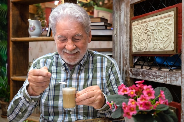 Porträt eines lächelnden älteren bärtigen Großvaters, der eine Tasse Kaffee mit Milch hält und einen Teelöffel Zucker setzt, glücklicher älterer Mann, der sich entspannt und die Pause mit heißem Cappuccino genießt