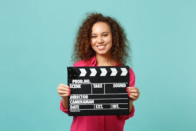 Porträt eines lächelnden afrikanischen Mädchens in Freizeitkleidung, das klassische schwarze Filmklappe hält, die auf blauem türkisfarbenem Hintergrund isoliert ist. Menschen aufrichtige Emotionen, Lifestyle-Konzept. Kopieren Sie Platz.