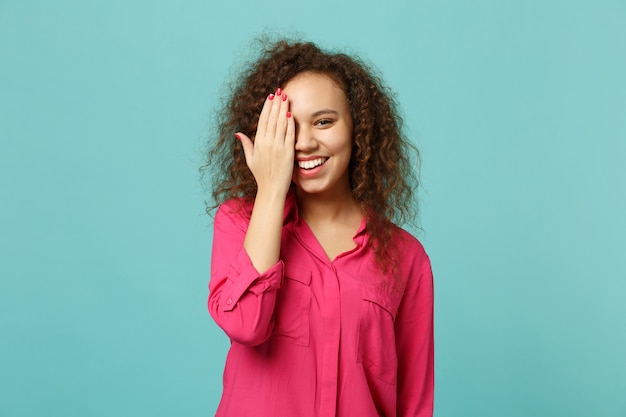 Porträt eines lächelnden afrikanischen Mädchens in rosafarbener Freizeitkleidung, das das Gesicht mit der Hand bedeckt, isoliert auf blauem türkisfarbenem Wandhintergrund im Studio. Menschen aufrichtige Emotionen, Lifestyle-Konzept. Kopieren Sie Platz.