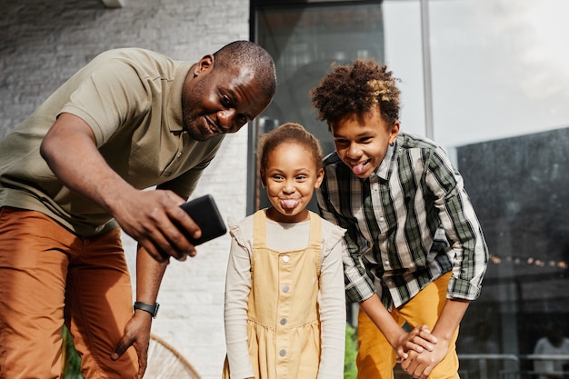 Porträt eines lächelnden afroamerikanischen Mannes, der ein lustiges Selfie mit zwei Kindern im Freien macht