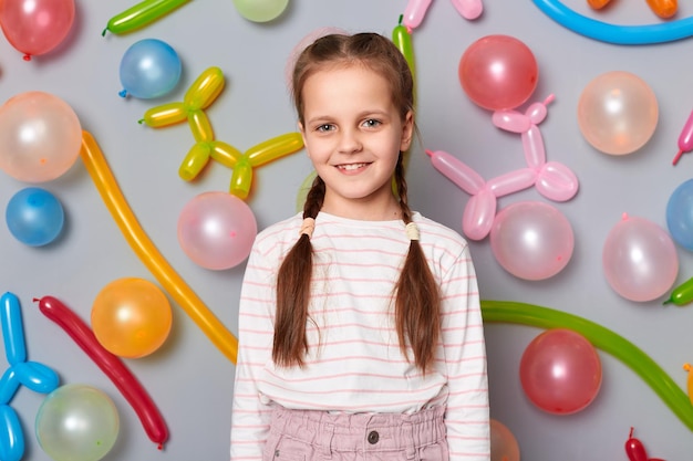 Porträt eines lächelnden, entzückenden, entzückenden kleinen Mädchens mit Zöpfen, das legere Kleidung trägt und isoliert vor grauem Hintergrund posiert, mit Luftballons, die mit glücklichem Gesicht, das Geburtstag feiert, in die Kamera blickt