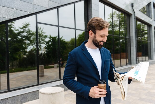 Porträt eines lächelnden Geschäftsmannes in Jacke, der Kaffee aus Pappbecher trinkt und Zeitung liest, während er im Freien in der Nähe des Gebäudes steht