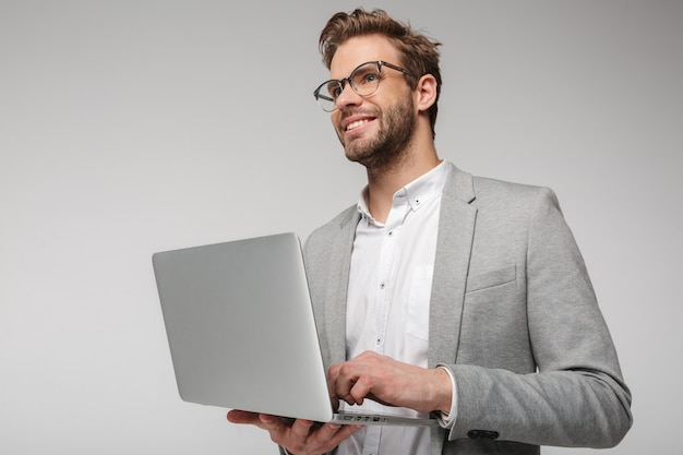 Porträt eines lächelnden gutaussehenden Mannes mit Brille, der Laptop isoliert über weißer Wand hält und benutzt