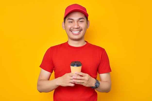 Porträt eines lächelnden jungen asiatischen Kuriermannes, der eine Kaffeetasse aus Papier hält und selbstbewusst isoliert über gelbem Hintergrund in die Kamera blickt