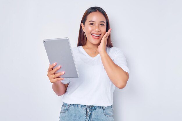 Porträt eines lächelnden jungen asiatischen Mädchens in weißem T-Shirt mit einem digitalen Tablet, das die Wange mit der Hand berührt und isoliert auf weißem Hintergrund in die Kamera blickt