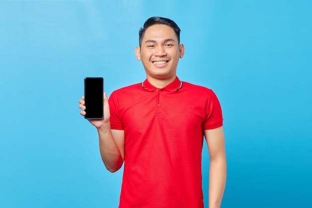 Porträt eines lächelnden jungen asiatischen Mannes, der ein Handy mit leerem Bildschirm zeigt und isoliert auf blauem Hintergrund in die Kamera blickt