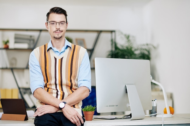 Foto porträt eines lächelnden jungen computeringenieurs mit brille, der am schreibtisch steht und in die kamera schaut