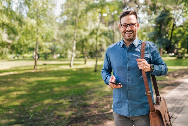 Porträt eines lächelnden jungen erwachsenen Mannes, der per Telefon durch den Park geht