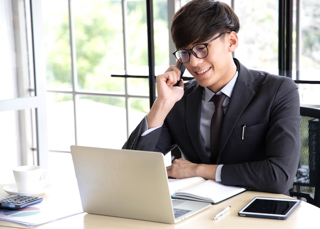 Porträt eines lächelnden jungen Geschäftsmannes, der auf seinem Smartphone spricht, während er an seinem Computer arbeitet und in seinem Büro am Schreibtisch sitzt, der einen schwarzen Anzug trägt.