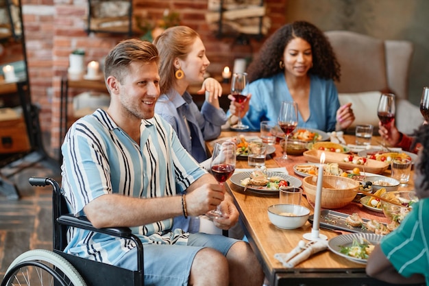 Porträt eines lächelnden jungen Mannes im Rollstuhl, der eine Dinnerparty mit Freunden genießt und ein Weinglas in der Hand hält