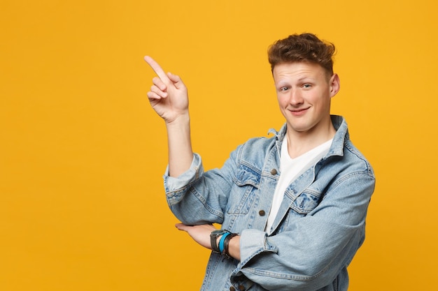 Porträt eines lächelnden jungen Mannes in Denim-Freizeitkleidung, der den Zeigefinger zur Seite zeigt, isoliert auf gelb-orangefarbenem Wandhintergrund im Studio. Menschen aufrichtige Emotionen, Lifestyle-Konzept. Mock-up-Kopienbereich.