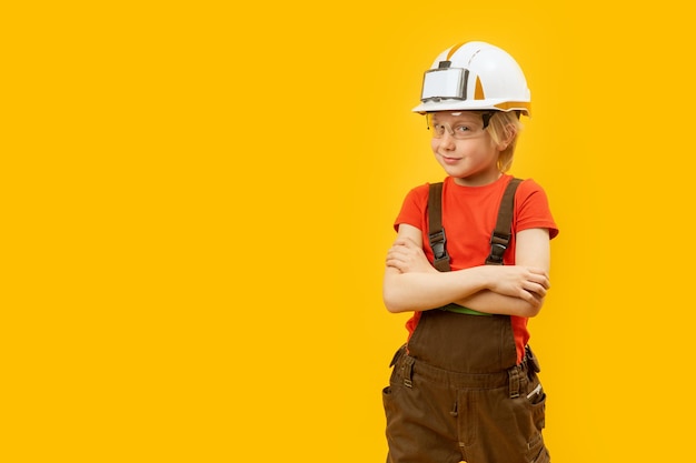 Porträt eines lächelnden Jungen mit Schutzhelm, Brille und Overall wie ein Arbeiter, gelber Hintergrund, Kopierraum-Attrappe