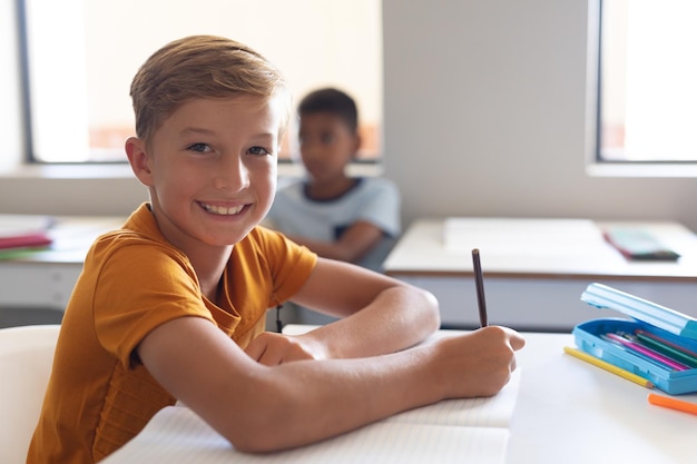 Porträt eines lächelnden kaukasischen Grundschülern, der im Unterricht am Schreibtisch sitzt und auf ein Buch schreibt