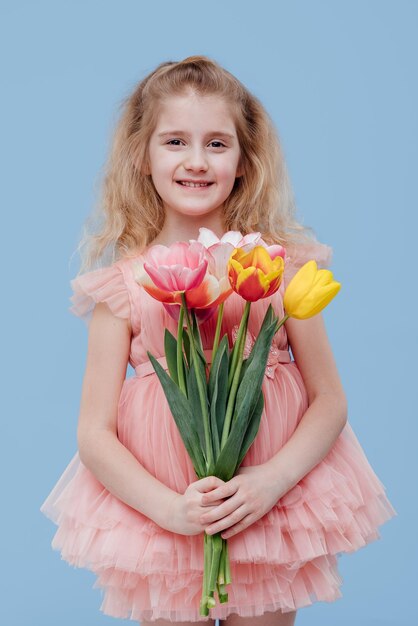 Porträt eines lächelnden kleinen Mädchens in rosafarbenem Kleid mit Frühlingsblumen in der Hand isoliert auf blauem Hintergrund