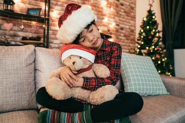 Porträt eines lächelnden kleinen Mädchens mit rotem Hut, das den Teddybär umarmt, der zu Hause auf der Couch sitzt. attraktives kind, das die augen schließt, schlief auf dem sofa ein, während es auf den weihnachtsmann wartete. winterwarmer Raum.