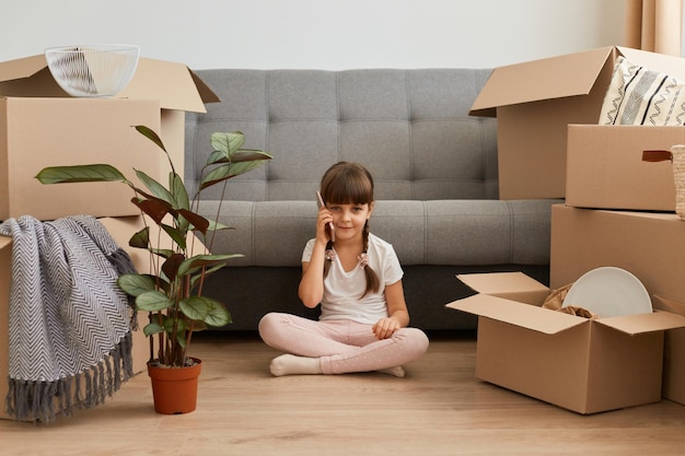 Porträt eines lächelnden kleinen mädchens mit zöpfen, das lässige kleidung trägt und auf einem boden in der nähe eines sofas und eines telefons sitzt und sich rühmt, dass sie mit ihrer familie in eine neue wohnung zieht.