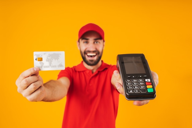 Porträt eines lächelnden Lieferboten in rotem T-Shirt und Mütze mit Kreditkarte und Zahlungsterminal isoliert über gelb