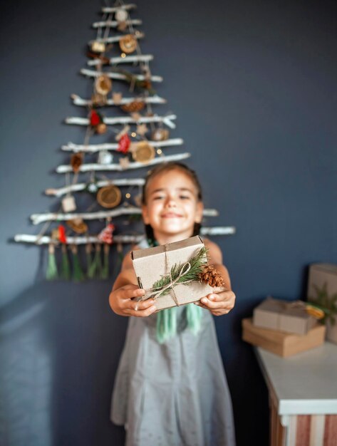 Foto porträt eines lächelnden mädchens, das ein geschenk gegen den an der wand hängenden weihnachtsbaum hält