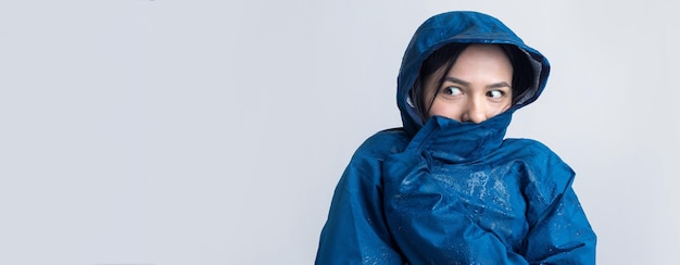 Porträt eines lächelnden Mädchens in blauem Regenmantel in Tropfen, das mit Kapuze auf grauem Hintergrund in einem Studio posiert