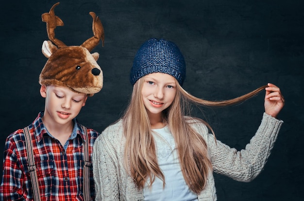 Porträt eines lächelnden Mädchens mit Wintermütze und süßer Junge mit Hirschmütze auf dunklem, strukturiertem Hintergrund.