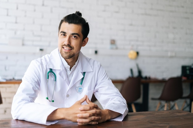 Porträt eines lächelnden männlichen Arztes in weißer Uniform mit Stethoskop, der am Schreibtisch im Krankenhausbüro sitzt und freundlich in die Kamera blickt