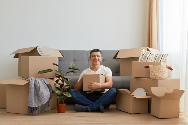 Porträt eines lächelnden, positiven Mannes mit weißem T-Shirt und Jeans, der mit Karton in den Händen auf dem Boden sitzt und glücklich ist, in eine neue Wohnung zu ziehen, um sein Glück beim Auspacken des persönlichen Stapels auszudrücken