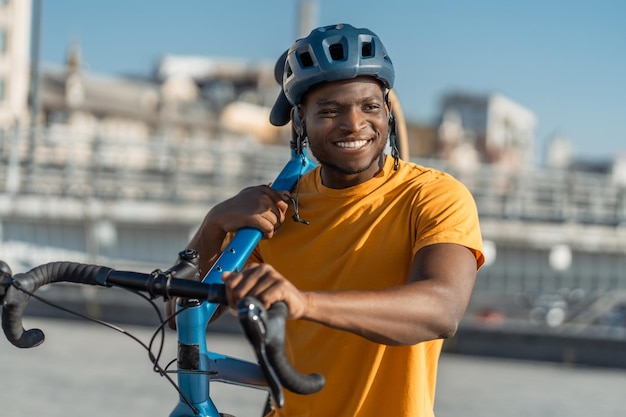 Porträt eines lächelnden, positiven nigerianischen Mannes, der ein Fahrrad hält und einen Schutzhelm auf einer städtischen Straße trägt