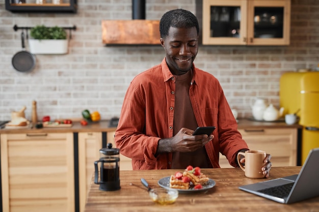 Porträt eines lächelnden schwarzen Mannes, der ein Smartphone benutzt, während er das Frühstück im gemütlichen Kopienraum der Küche zu Hause genießt