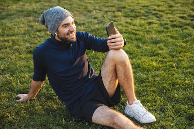 Porträt eines lächelnden sportlichen Mannes, der im Trainingsanzug mit Smartphone gekleidet ist und während des Trainings im grünen Park auf Gras sitzt