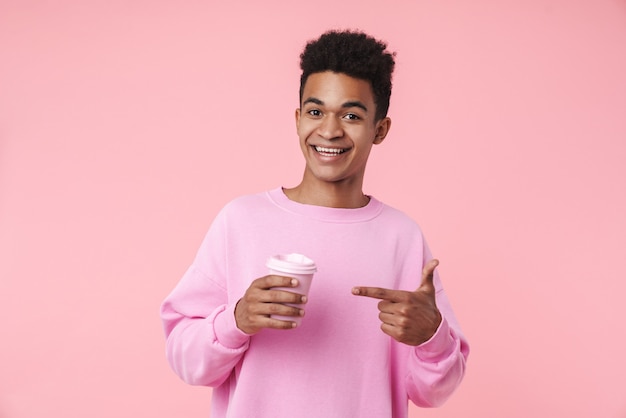 Porträt eines lächelnden Teenager-Jungen mit Pullower, der isoliert über rosa Wand steht, Kaffeetasse zum Mitnehmen hält und mit dem Finger zeigt