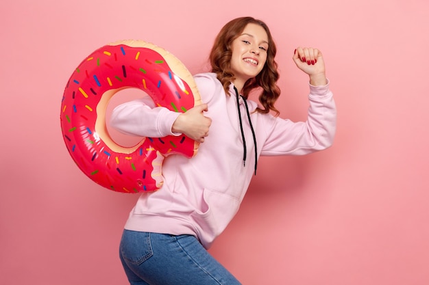 Porträt eines lächelnden Teenager-Mädchens mit lockigem Haar in Hoodie, das mit einem rosafarbenen Donut mit Gummiring spazieren geht, bereit für den Urlaub. Studioaufnahme im Innenbereich isoliert auf rosa Hintergrund