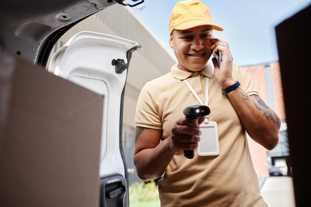 Porträt eines lächelnden Zustellers, der per Smartphone spricht und Codes auf Paketen im Lieferwagen scannt