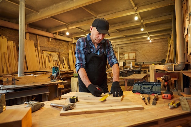 Porträt eines leitenden Tischlers, der mit einem Bleistift mit einer Holzmarkierungsplanke arbeitet und Messungen vornimmt, um ein Stück Holz zu schneiden, um ein Möbelstück in der Tischlerei herzustellen