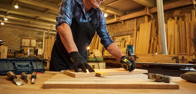 Foto porträt eines leitenden tischlers, der mit einem bleistift mit einer holzmarkierungsplanke arbeitet und messungen vornimmt, um ein stück holz zu schneiden, um ein möbelstück in der tischlerei herzustellen
