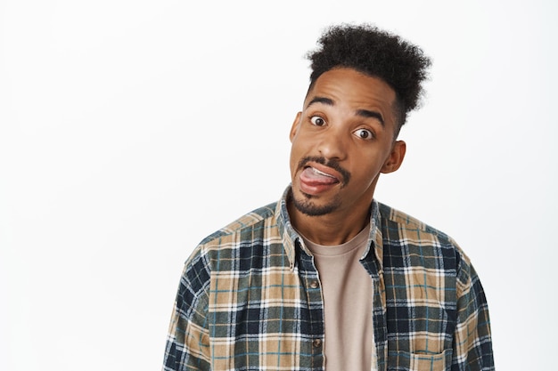 Porträt eines lustigen afroamerikanischen Mannes, der dumme Gesichter, Zunge und Grimasse vor der Kamera zeigt, Spaß hat und in einem lässigen, karierten Hemd auf weißem Hintergrund steht
